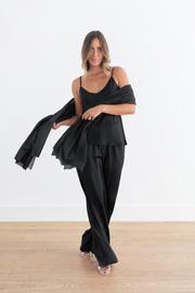 A woman modeling a black two-piece outfit consisting of a spaghetti-strap camisole top and matching wide-leg trousers, holding a matching black sheer wrap or shawl against a white wall and light wood floor.